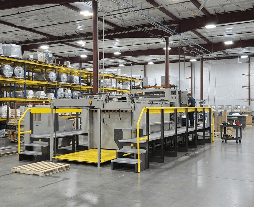 A large industrial machine with yellow safety rails sits in a spacious warehouse with shelves of supplies, pallets, and a worker nearby. The environment is well-lit with exposed ceiling beams.